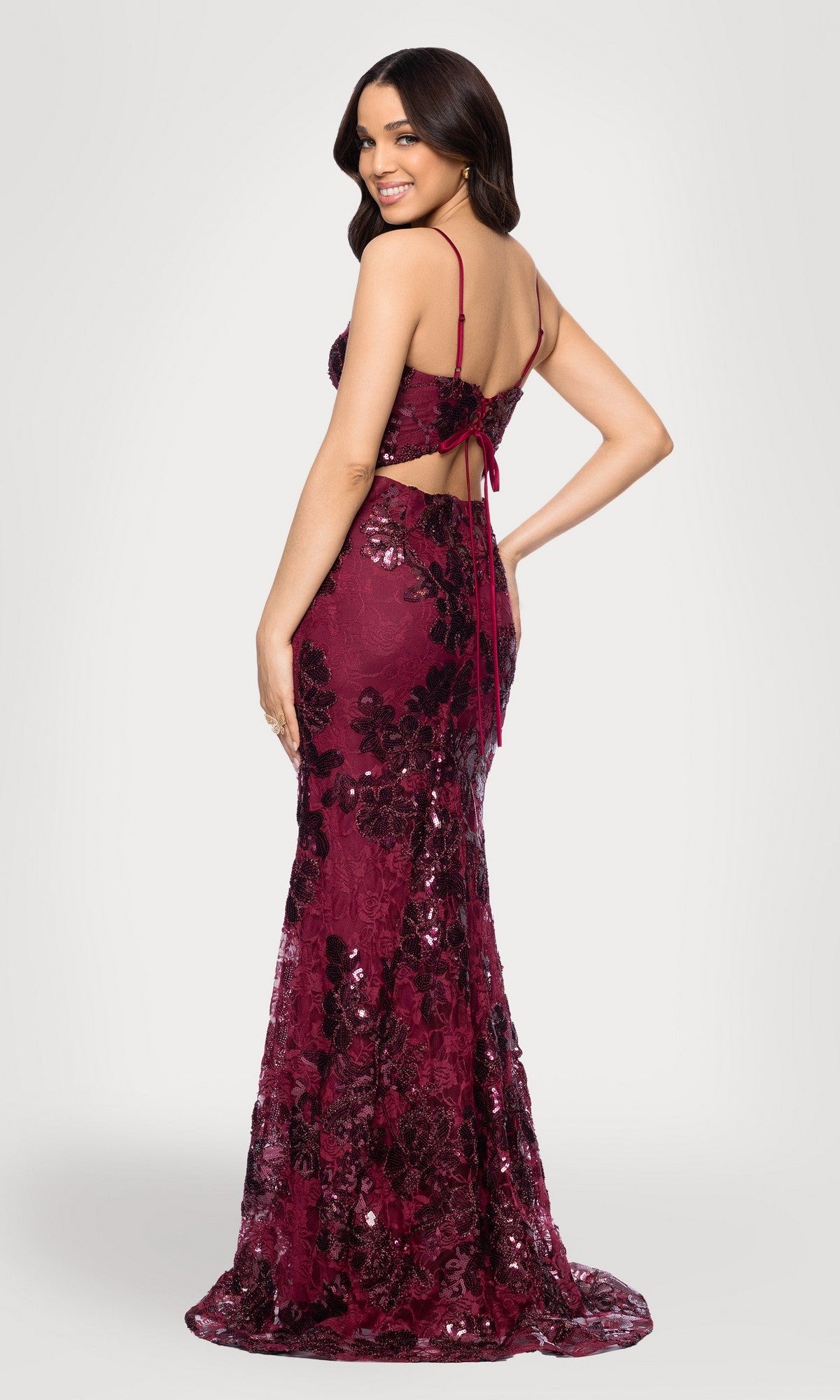 A woman smiles over her shoulder wearing the Blondie Nites 5883BN, a burgundy floor-length prom dress with fitted sequin lace, spaghetti straps, floral pattern, and an open back.